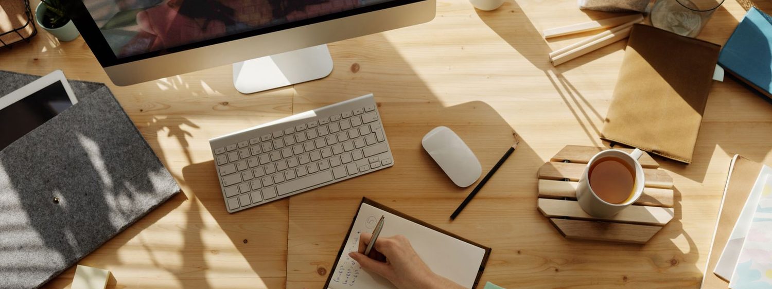 Escritorio de trabajo iluminado con un computador, un cuaderno y una taza de té mientras alguien toma notas